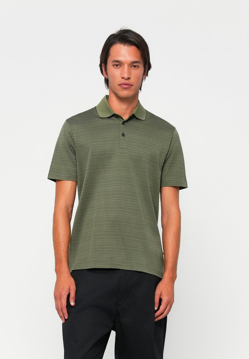Kurzärmliges Poloshirt in Olivgrün mit strukturiertem Design, weichem Kragen und einer Knopfleiste mit drei Knöpfen. Kombiniert mit dunklen Hosen.