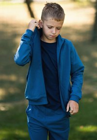 Niebieska bluza z zamkiem wykonana z miękkiego materiału, noszona z ciemnym t-shirtem pod spodem. Bluza ma ściągacze na rękawach i luźny krój.