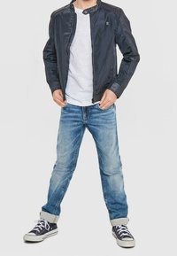 Pojke iklädd en svart zip-up jacka, vit t-shirt, uppvikta blå jeans och svarta sneakers med vita snörningar, står med händerna i fickorna.