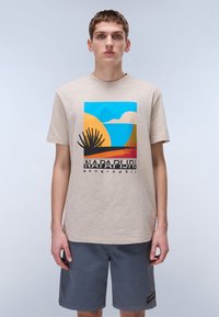 Bež bombažno majico z barvitim grafičnim motivom pokrajine s silhuetami palm. V kombinaciji s sivo kratkimi hlačami, nevtralna ozadje.