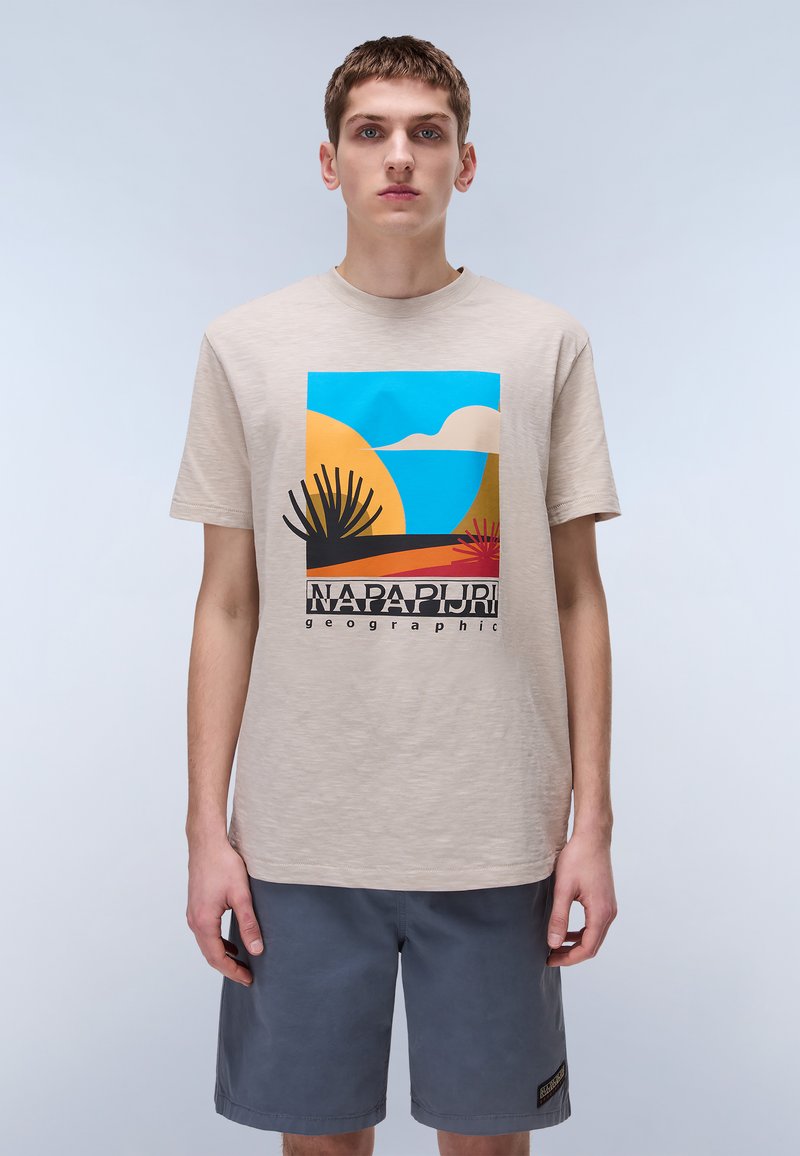Bež bombažno majico z barvitim grafičnim motivom pokrajine s silhuetami palm. V kombinaciji s sivo kratkimi hlačami, nevtralna ozadje.