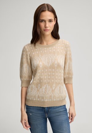 Młoda kobieta o brązowych włosach, ubrana w beżowy sweter z wzorem, z rękawami do łokci oraz niebieskie dżinsy, na jednolitym tle.