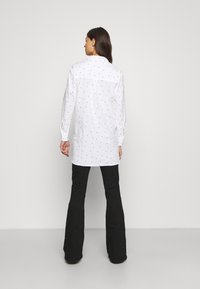 Camisa de manga comprida branca com bolinhas pretas, com botão frontal e uma yoke nas costas. Combinada com calças pretas largas. Textura de tecido suave.