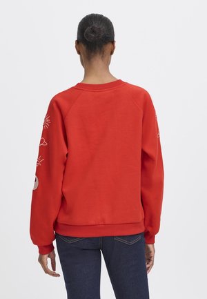 Mujer con el pelo oscuro recogido en un moño, lleva una sudadera roja con diseños gráficos blancos en las mangas y vaqueros azul oscuro, de espaldas.