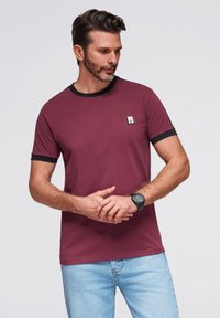 Burgundské bavlněné tričko s černým lemem na límci a rukávech; obsahuje malé logo značky na hrudi. Nosené s světle modrými džíny.