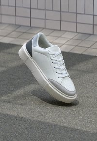 Sneaker bianco con accenti grigi e neri, lacci bianchi e suola spessa, posizionato sopra uno sfondo in pavimentazione testurizzata e parete piastrellata.