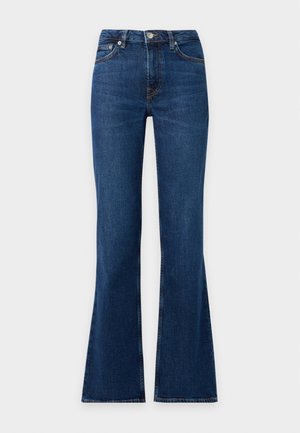 Mujer de pie con chaleco de punto gris sobre camisa blanca, vaqueros azules, mocasines oscuros, sosteniendo un pequeño bolso negro en la mano derecha.