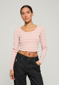 Rosa und lila gestreiftes, halb langärmeliges Oberteil mit einem gewellten Saum und Knopfverschluss, kombiniert mit hochgeschnittenen schwarzen Jeans.