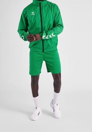 Man draagt een groen sportjack en korte broek met witte sneakers en sokken, staand tegen een effen lichte achtergrond.