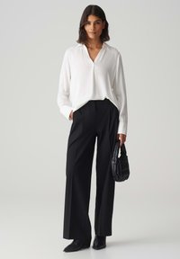 Weiße Bluse mit langen Ärmeln und V-Ausschnitt, kombiniert mit hochgeschnittenen schwarzen Hosen. Schwarze strukturiert Handtasche und schwarze spitze Stiefel.
