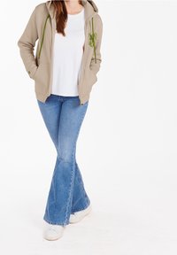 Felpa beige con zip e coulisse verdi, indossata sopra una t-shirt bianca, abbinata a jeans svasati blu e sneakers bianche.