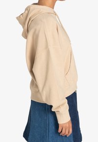 Beige hættetrøje med lomme foran, faldede skuldre og ribbede ærmeafslutninger, kombineret med et denimskørt. Blød tekstur.