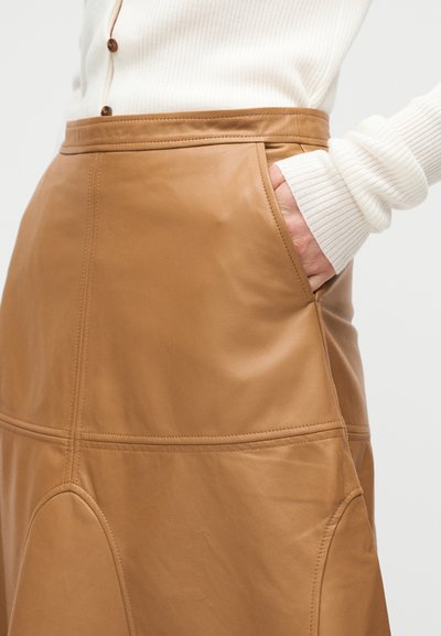 Γυναίκα που φοράει καφέ δερμάτινη φούστα με το χέρι στην τσέπη, σε συνδυασμό με λευκό πλεκτό πουλόβερ με καφέ κουμπιά.