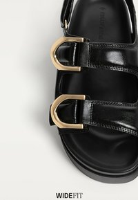 Sort lædersandal med to guldtonede spænder, åben tå-design og en glat tekstur. Tykt sål for stabilitet og støtte.