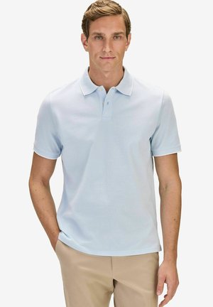 Mann trägt ein hellblaues kurzärmeliges Polohemd und beige Hose, steht mit einer Hand in der Tasche vor einem einfarbigen Hintergrund.