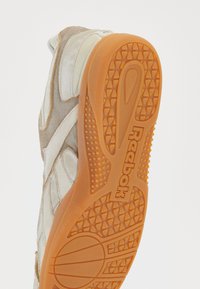 Tenis Reebok care prezintă o combinație de material textil bej și piele întoarsă, având o talpă din cauciuc portocalie. Talpa are un model texturat pentru aderență.