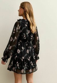 Vrouw met lang blond haar, gekleed in een zwarte bloemenjurk met doorschijnende lange mouwen, ruches en een tailleceintuur, kijkt naar links tegen een effen muur.