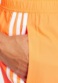 Pantalones cortos deportivos naranjas hechos de tela ligera, con franjas blancas y un bolsillo lateral. Textura suave y cinturilla elástica.