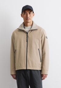 Beige Zip-Jacke mit hohem Kragen, Reißverschlusstaschen und weicher Textur. Getragen über einem hellen Hemd, kombiniert mit dunklen Hosen.