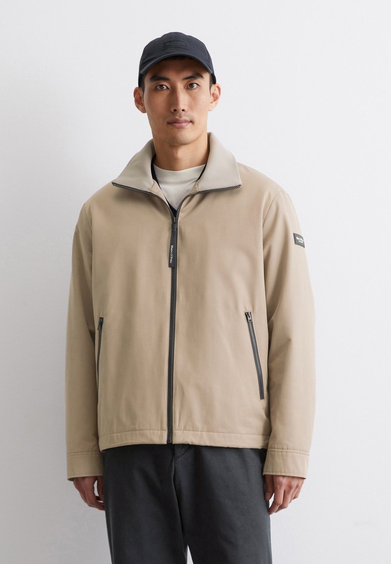 Beige Zip-Jacke mit hohem Kragen, Reißverschlusstaschen und weicher Textur. Getragen über einem hellen Hemd, kombiniert mit dunklen Hosen.