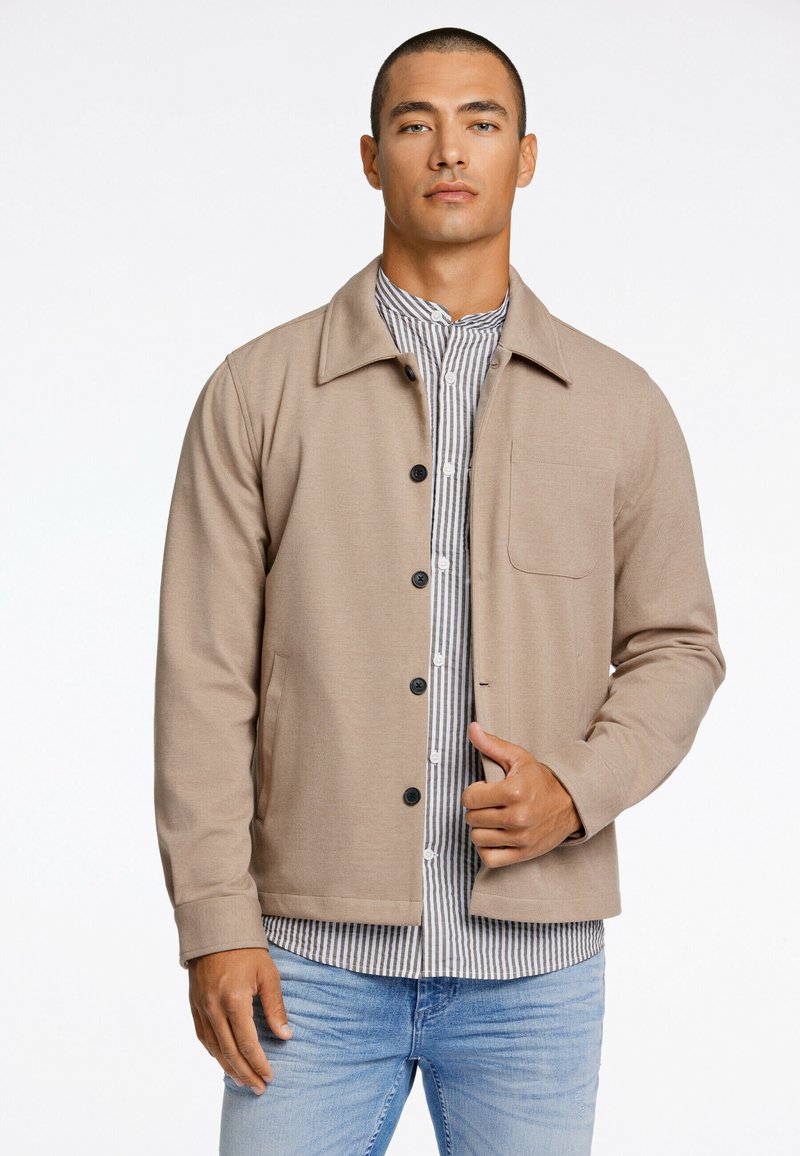 Beige Jacke mit Kragen, Knopfleiste und einer einzelnen Brusttasche, über einem gestreiften Hemd getragen, kombiniert mit hellblauen Jeans.