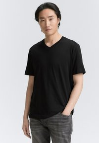 Schwarzes V-Ausschnitt-T-Shirt aus weichem Stoff, mit kurzen Ärmeln und lockerer Passform. Das Model trägt graue Jeans in einer lässigen, mit Taschen versehenen Haltung.