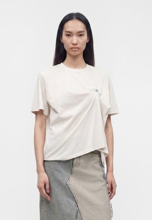 Jeune femme aux longs cheveux foncés porte un t-shirt ample blanc cassé et une jupe en jean grise bicolore, debout devant un fond clair uni.