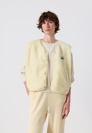 Persoon met kort krullend haar die een crèmekleurig fleecevest draagt over een lichte trui en getextureerde broek tegen een effen witte achtergrond.