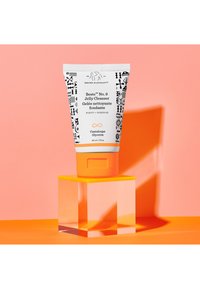 Jelly cleanser tube features a white background with black patterns, orange cap, and accents. It rests on a clear block against a pink backdrop.