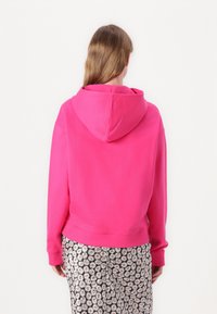 Felpa con cappuccio rosa, con maniche lunghe, tasca a marsupio e polsini a costine. Indossata sopra pantaloni a fantasia floreale bianco e nero.