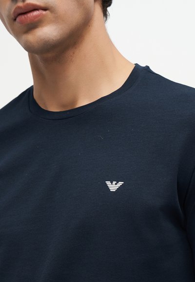 Gros plan d'une personne portant un t-shirt bleu marine foncé avec un petit logo d'aigle stylisé blanc sur la poitrine.