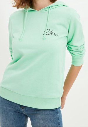 Frau trägt einen hellgrünen Hoodie mit dem Wort „Slow“ auf der Brust, kombiniert mit blauen Jeans.