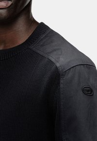 Gros plan sur une personne à la peau foncée portant un pull noir à côtes avec des empiècements en tissu lisse sur les épaules et un logo en relief sur la manche.