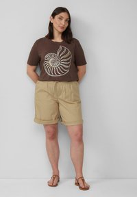 Braunes Grafik-T-Shirt mit einem weißen Spiraldesign, kombiniert mit beigen, hochgerollten Shorts und braunen flachen Sandalen, was einen lässigen Sommerlook unterstreicht.