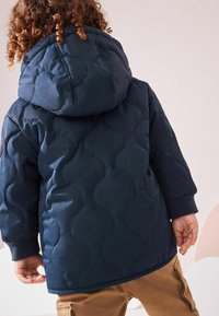 Marinblå quiltad pufferjacka med huva, som har en vågig textur och ribbade manschetter. Avslappnad passform, kombinerad med khakifärgade byxor.