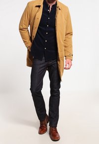 Kaki trenchcoat över en mörkblå skjorta med knapp, tillsammans med mörka jeans och bruna läderstövlar. Kappan har krage och knappar.