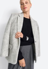 Hellegrauer Filzmantel mit Reverskragen, zwei vorderen Taschen und einem Knopfverschluss, getragen über einem schwarzen Strickpullover und grauen Hosen.