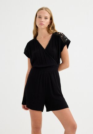 Mujer joven con cabello largo y rubio, vistiendo un mono negro con detalles de encaje en los hombros, posando frente a un fondo blanco liso.