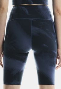Primo piano di una persona che indossa pantaloni da ciclismo a vita alta, blu scuro tie-dye, in piedi con le braccia rilassate lungo i fianchi.