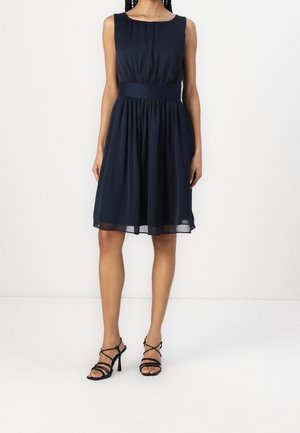 Femme portant une robe bleu marine sans manches longueur genou avec une taille ajustée et des sandales noires à talons hauts et brides, debout sur un fond blanc.
