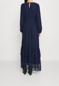 Granatowa, długa sukienka maxi z długim rękawem, z falbanowym dołem, fakturą w kropki i ozdobnym otworem na plecach. Wykończona paskiem w talii.