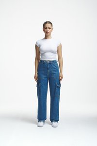 Young woman standing facing forward wearing a white fitted t-shirt, high-waisted blue cargo jeans, and white sneakers on a white background.