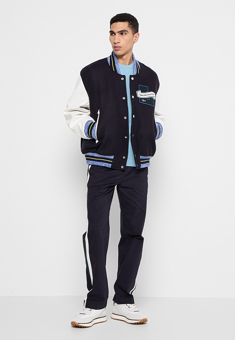 Veste varsity bleu marine et crème avec des accents rayés bleus, boutons-pression et un patch. Portée sur un pull bleu clair et un pantalon sombre.