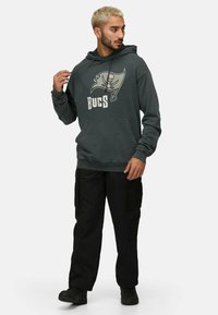 Felpa grigia con logo dei Buccaneers e la scritta "BUCS," abbinata a pantaloni cargo neri. La felpa ha una tasca frontale e cappuccio con cordini.