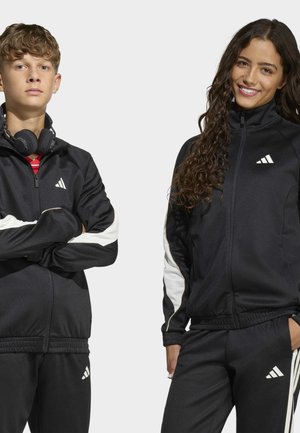 Tienerjongen en -meisje dragen zwarte Adidas trainingspakken, de jongen met een koptelefoon om zijn nek en met gekruiste armen, het meisje glimlacht met één hand in de zak.