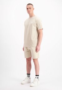 Beige t-shirt met korte mouwen met een klein logo op de borst, gecombineerd met bijpassende shorts, witte sneakers en zwart-wit gestreepte sokken.