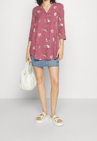 Rozenpatroon, lange mouwen tuniek top met een V-hals, gedragen over een denim mini-rok en gecombineerd met witte sandalen en een crèmekleurige handtas.