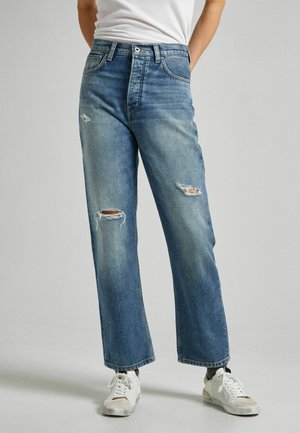 Jeans de denim azul claro con detalles desgastados, corte recto y cintura alta. Presenta un cierre de cremallera y cinco bolsillos. Combinados con zapatillas blancas.