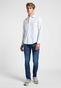 Camisa blanca de manga larga con botones combinada con jeans azules. La camisa tiene un logo sutil y un cuello clásico. El calzado son zapatillas grises.