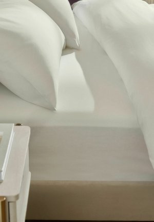 Bett mit zwei weißen Kissen und weißen Laken neben einem beigen Nachttisch mit Büchern und weichem natürlichem Licht.